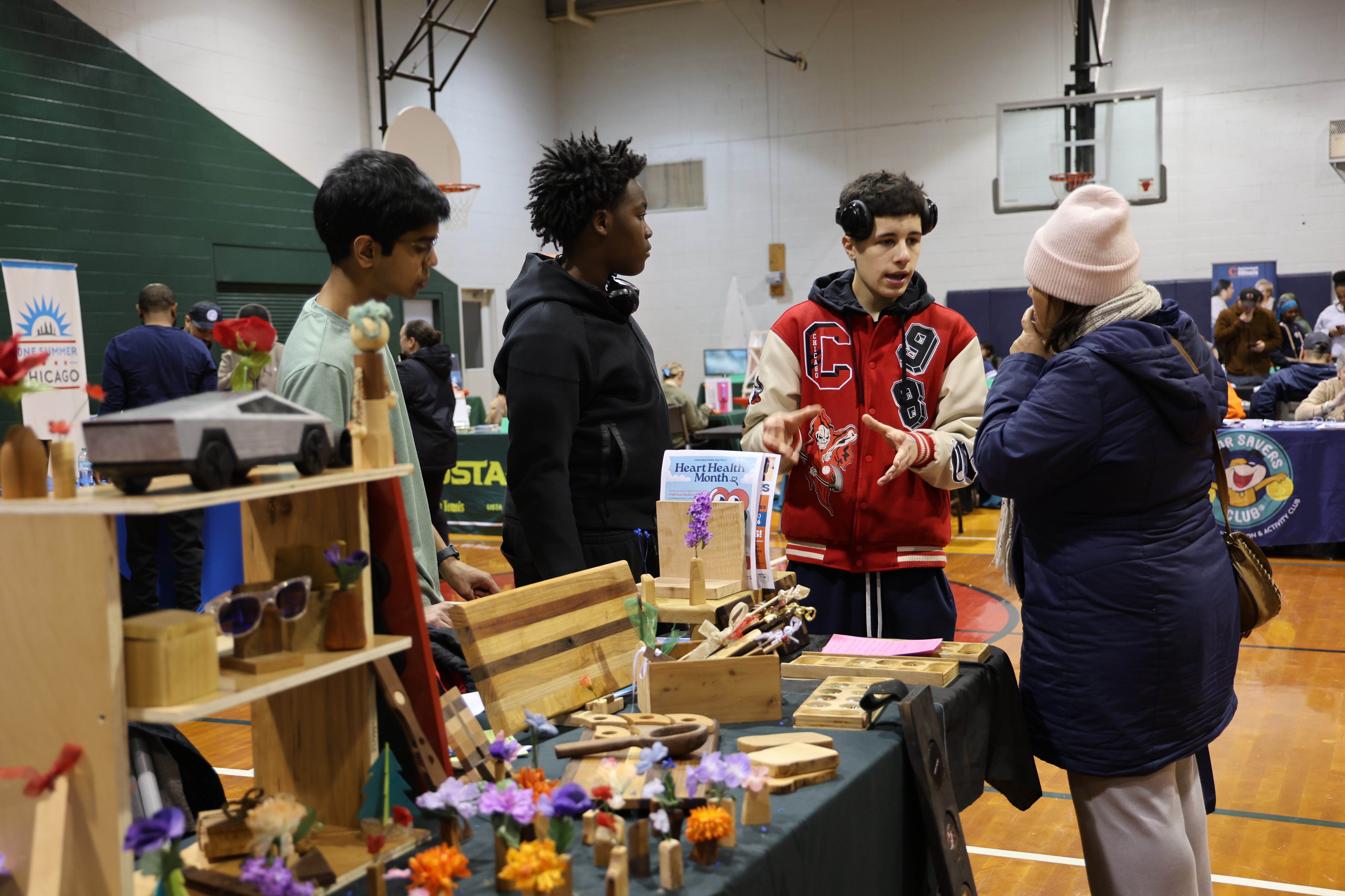 Youth in a varsity jacket talks at a craft table with handmade items in a gymnasium.